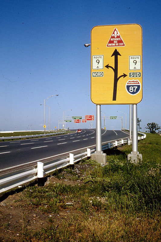 La Prairie. <br><br>
<i>These are still the old Quebec shields, before the grand renumbering. What is now Autoroute 10, was still the Autoroute des Cantons de L’est, and its shield was a red triangle. What really fascinated me was that the guide signs specific to that route in the distance were also red. I know that other toll autoroutes had a similar triangular shield in different colors, including blue and green (BGS, BBS and BRS – sheesh!). Would those guide signs be matched to the toll road color? Of course an Interstate shield in Canada is always something special.</i>